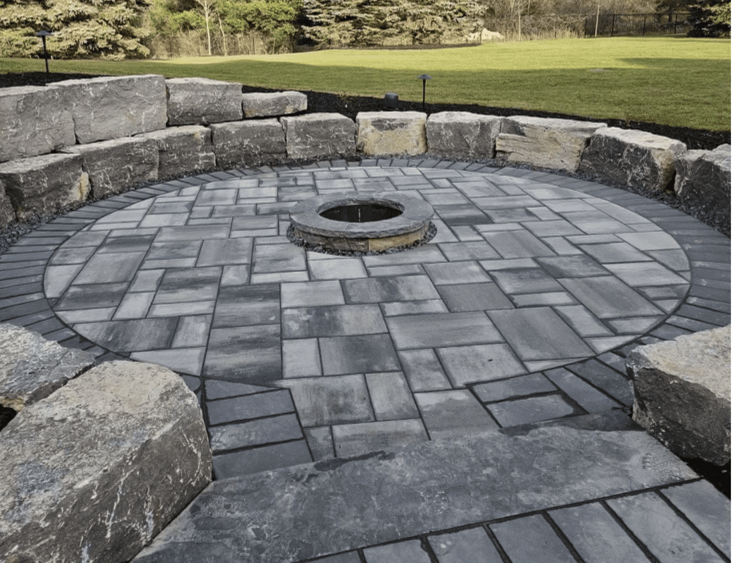 Guelph ON home with large armour stone retaining wall surrounding a round exterior fireplace area, featuring grey pavers and charcoal border.